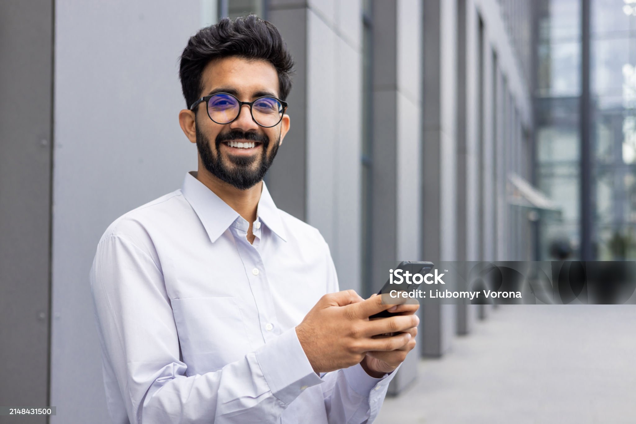 Portrait of a smiling and happy young Indian man in a white shirt and shorts standing near an office center, holding a phone and smiling at the camera.